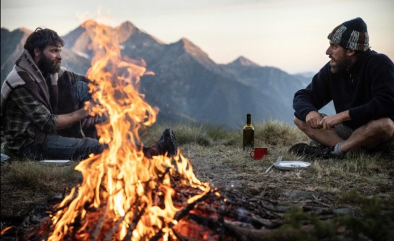 Two men sit by a bonfire with mountains in the background